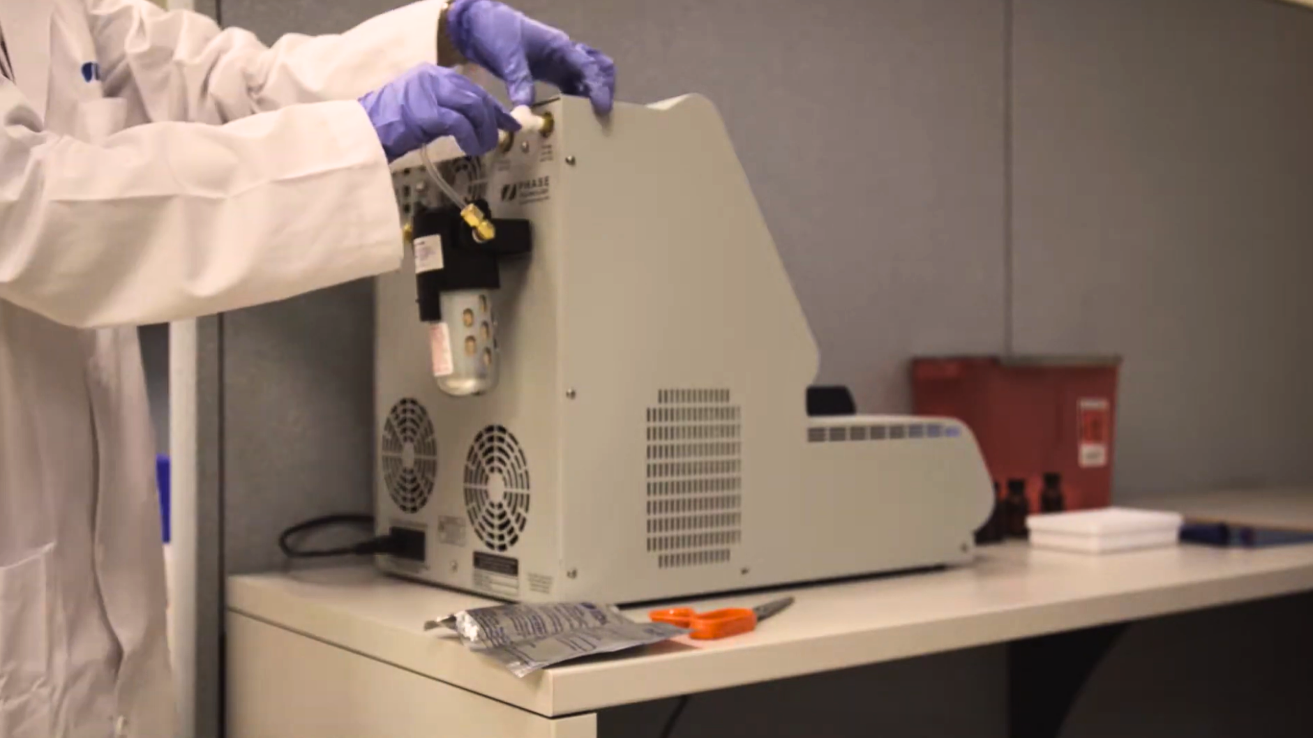 A lab technician in a white coat and blue gloves conducts maintenance on a JFA-70Xi analyzer on a table. Nearby are tools and a red container. The scene conveys a professional lab environment.