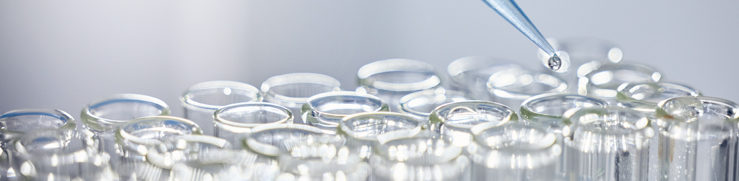 A blue pipette dripping liquid into a row of clear glass test tubes. The image conveys a scientific and precise atmosphere in a leak testing laboratory setting.