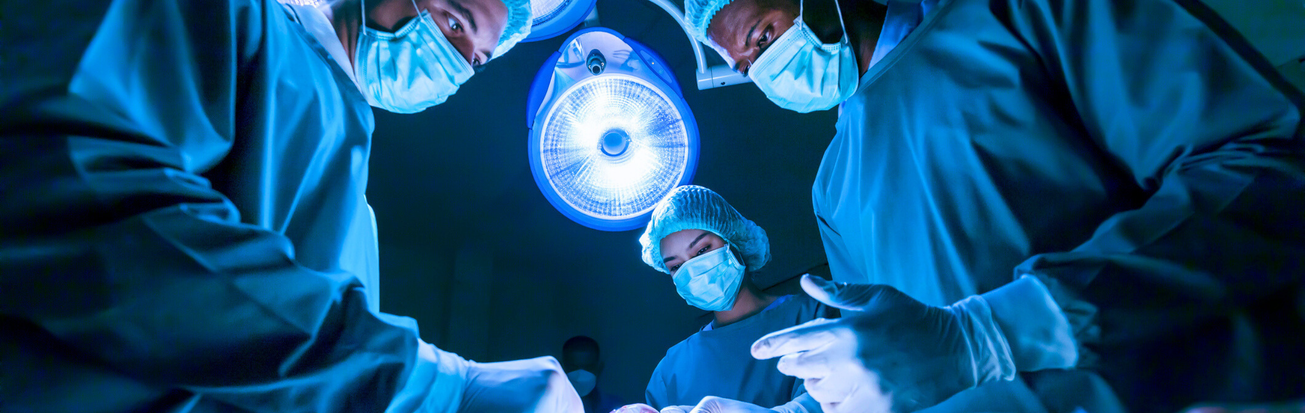 Three surgeons in blue scrubs and masks perform a surgical procedure under a bright operating room light.