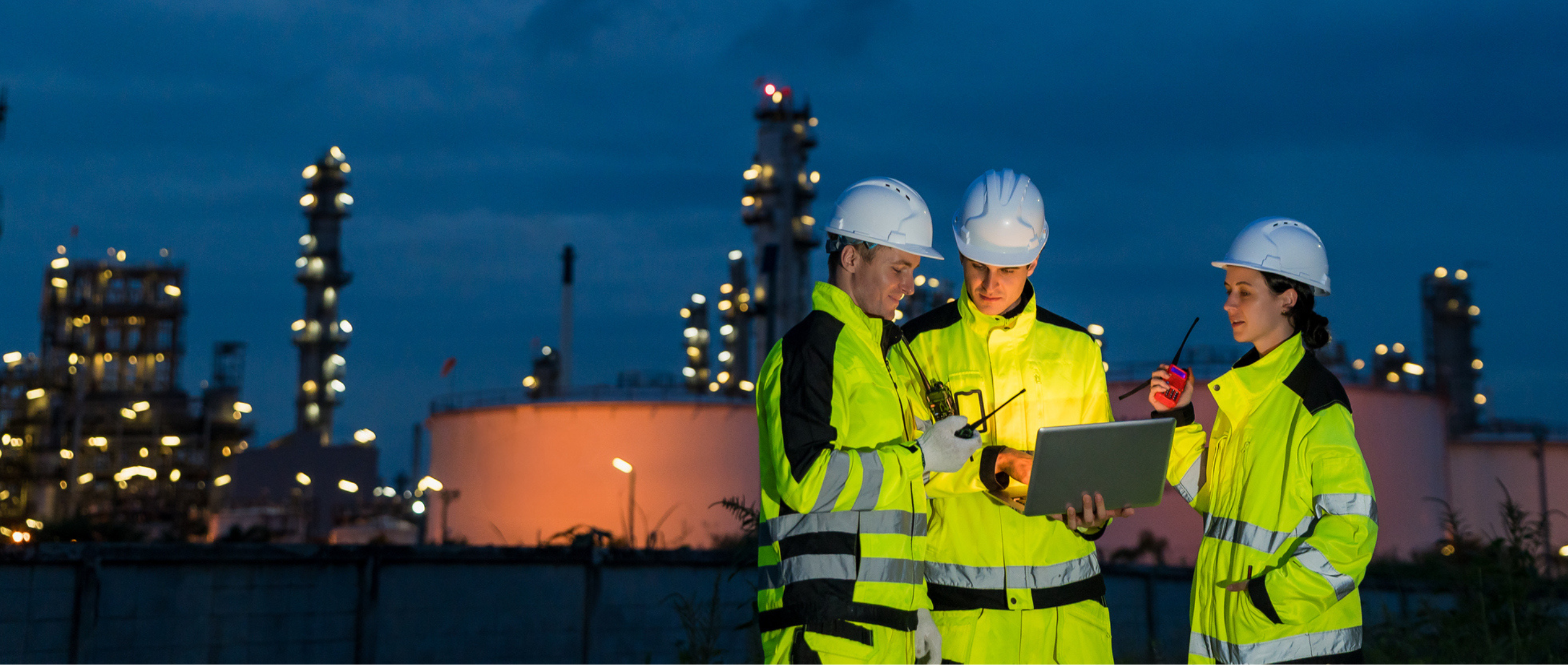 Three workers in reflective jackets and helmets discuss the news using a laptop at a refinery at night. The illuminated plant glows in the background.