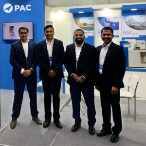 Four men in suits stand in a corporate booth with PAC branding. They are smiling, conveying a professional and welcoming tone.