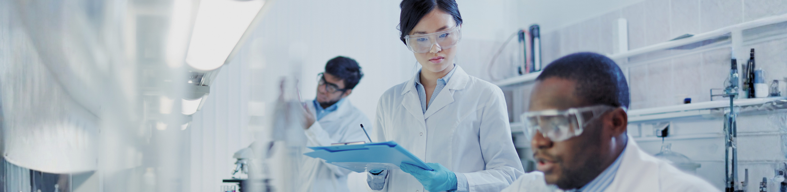 Three scientists in a lab, wearing protective goggles and white coats, conducting experiments. A focused, collaborative atmosphere is depicted. Header for About Us webpage.