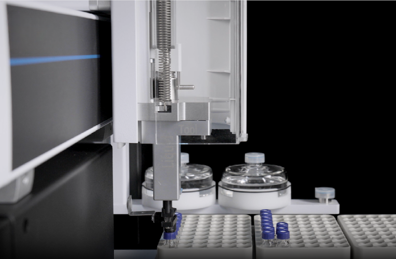 A robotic arm in a laboratory setting places a vial on a tray. Nearby, there are stacks of additional vials and two transparent circular containers. Image for elemental analysis and chromatography pages.