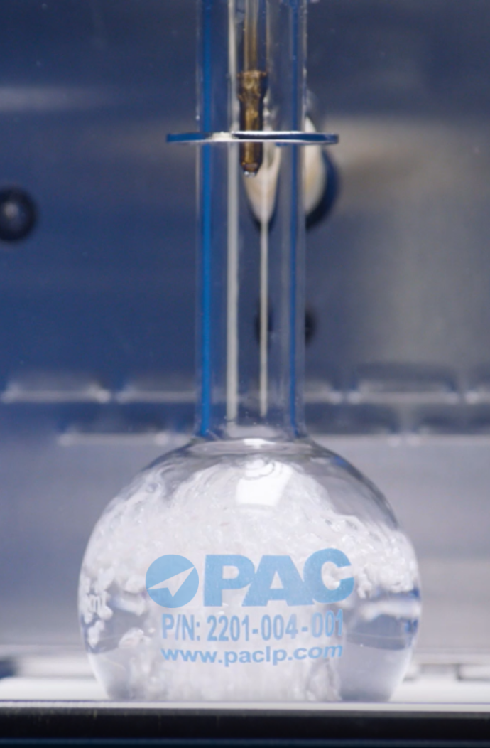 A laboratory flask with a narrow neck and bulbous bottom is placed in a blue apparatus for distillation testing. The flask contains a foamy white liquid. The logo 
