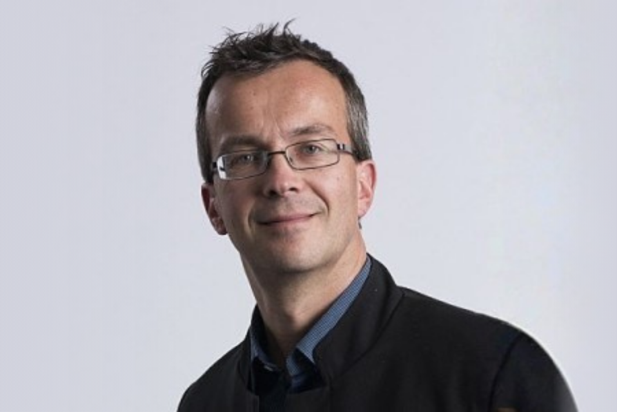 Smiling man with short brown hair and glasses wearing a dark jacket over a shirt, standing against a plain white background, conveying a friendly demeanor.