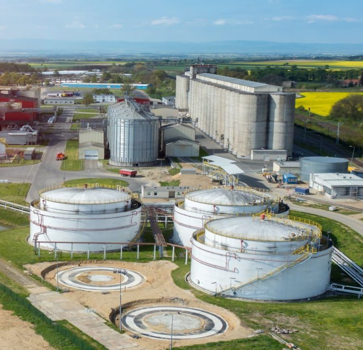 Aerial view of an industrial complex with large storage tanks and smokestacks emitting smoke. Surrounded by fields, the scene feels expansive and industrious.