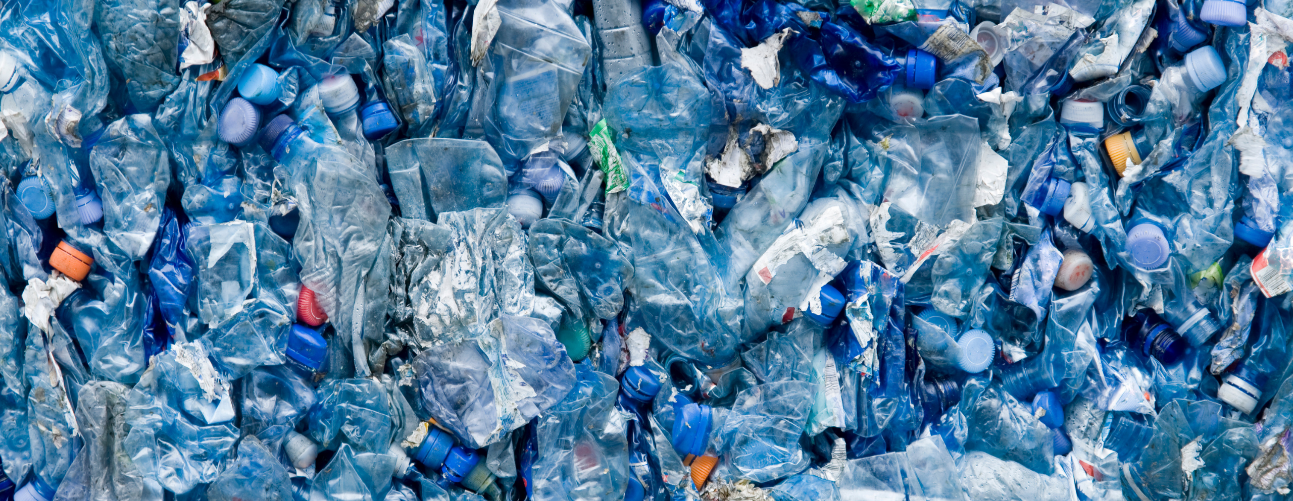 A densely packed pile of crushed, empty plastic bottles in various shades of blue, with some colorful caps visible. The image conveys the waste plastic pyrolysis process.