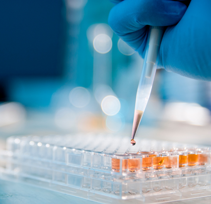 A gloved hand holds a pipette over a transparent tray with orange liquid, in a lab setting. The scene conveys precision and a scientific focus.