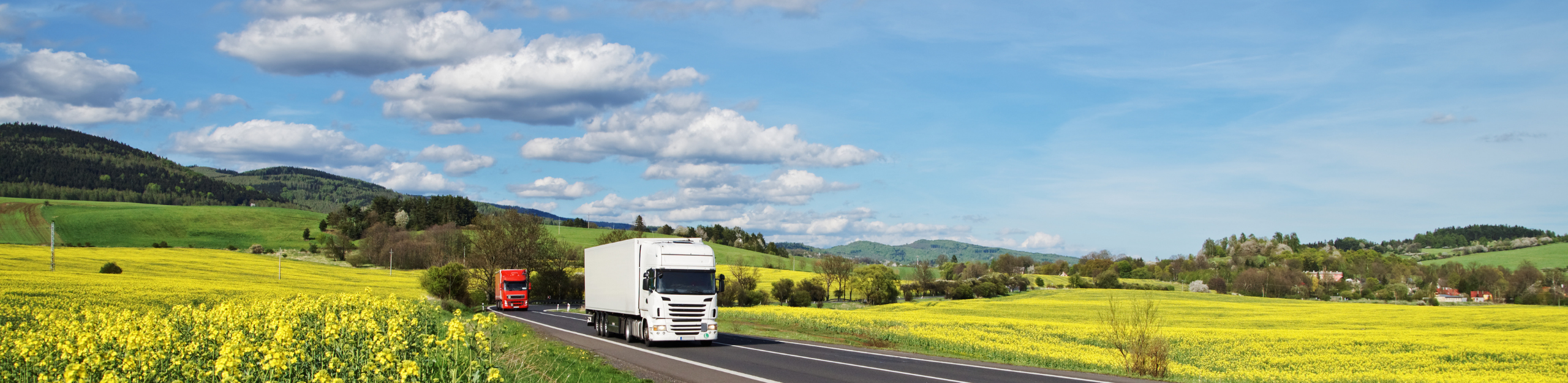 A white truck drives on a rural road bordered by vibrant yellow fields and green hills under a bright blue sky with scattered clouds, intended to depict renewable fuels testing.