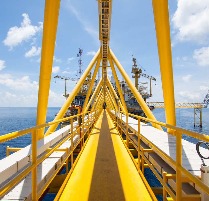 A bright yellow bridge leads to an offshore oil rig under a sunny sky. The structure reflects on the calm, blue sea, evoking a sense of industrial might.