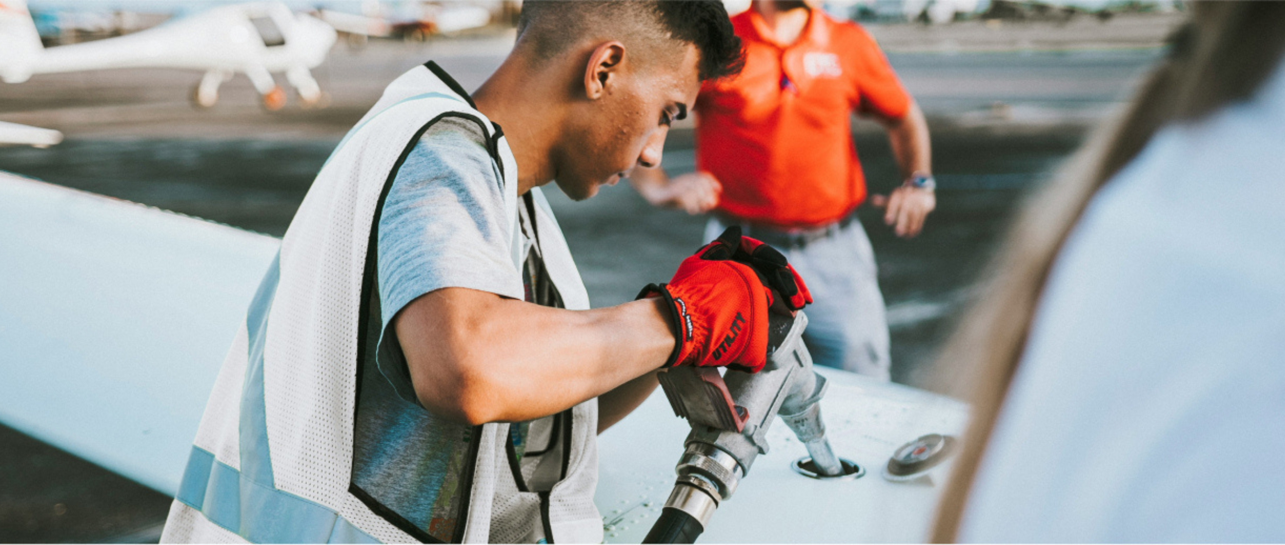 A focused worker in a reflective vest and red gloves fuels an aircraft. Another person in a red shirt stands nearby, set against an airport backdrop.