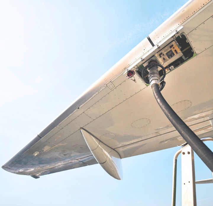 A plane's wing is captured from underneath against a clear blue sky, with a fueling hose connected to its underside, highlighting technical precision. PAC Powered by Emcee.