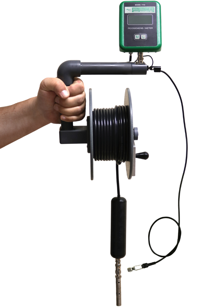 A hand holds a green PAC Powered by Emcee model 1152 Digital Conductivity Meter and a black cord reel connected to a sensor probe against a black background, suggesting technical measurement.