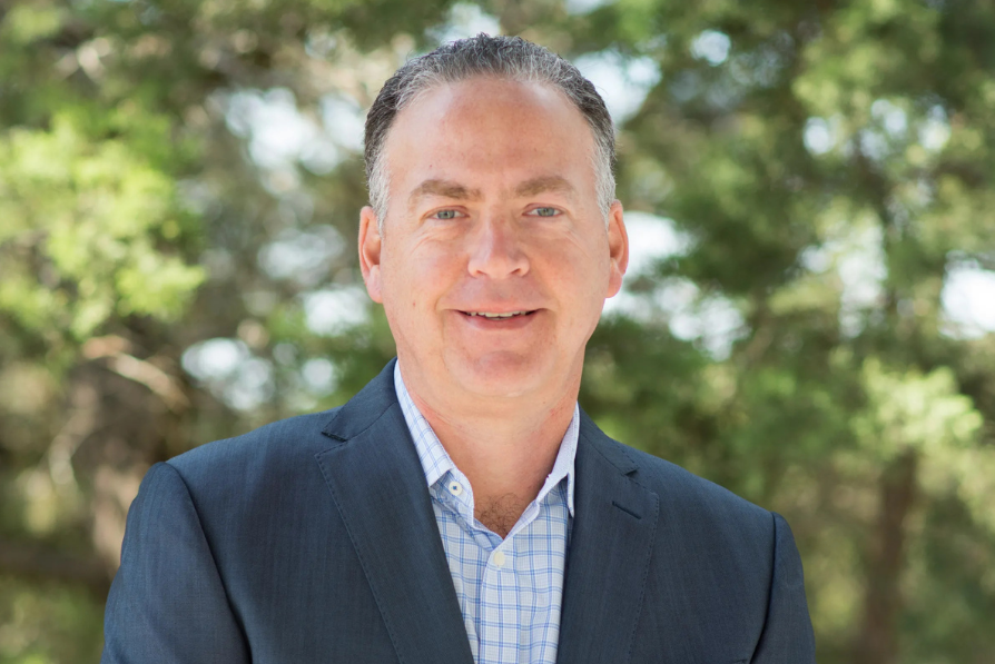Smiling man in a suit stands outdoors with blurred green trees in the background, conveying a professional and approachable vibe.