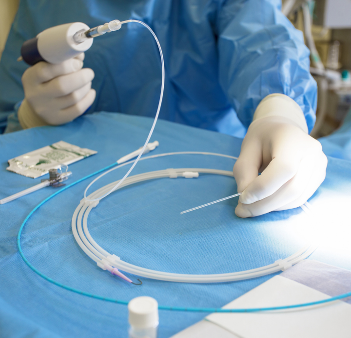A medical professional in blue scrubs and white gloves holds a catheter and surgical instrument over a sterile table, conveying precision and focus.