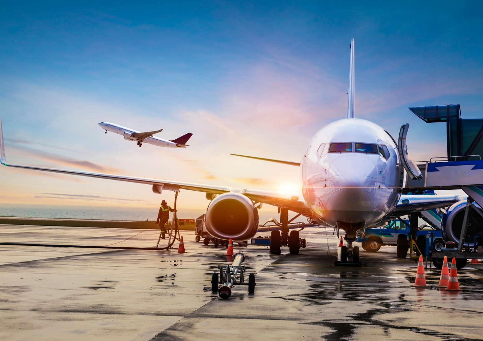 A commercial airplane is parked at an airport gate under a vibrant sunset. Another plane takes off in the background. The scene conveys travel and adventure. SAF - Sustainable Aviation Fuel.