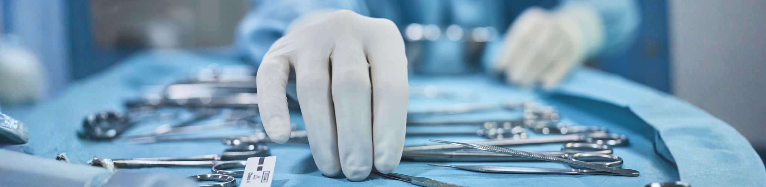 A gloved hand reaches for surgical instruments on a blue sterile tray in an operating room, conveying precision and focus. Uson.