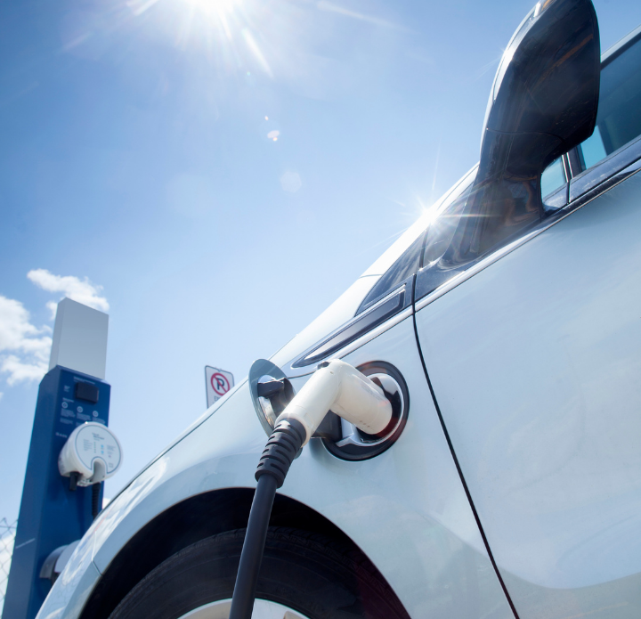 Electric car charging at a station under a bright blue sky. The white vehicle is plugged in, with sunlight reflecting off its surface, conveying a sense of innovation.