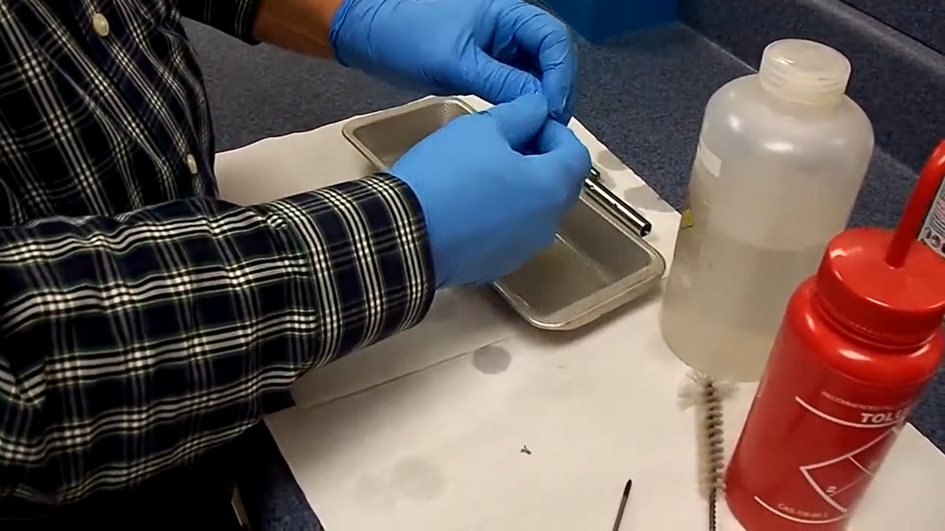 A person in a plaid shirt and blue gloves is cleaning a small item over a tray on a counter. Nearby are bottles, a brush, and a white cloth.