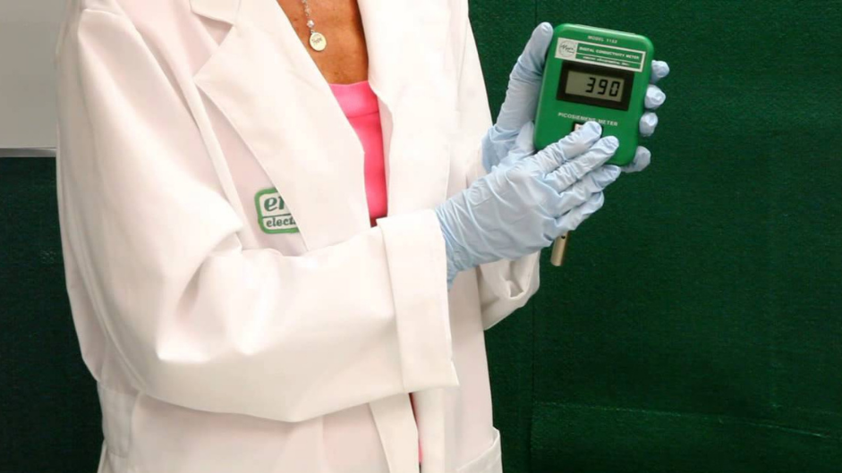 A person in a lab coat and blue gloves holds a green electronic device with a display screen. A black case is on the table, suggesting a scientific setting.