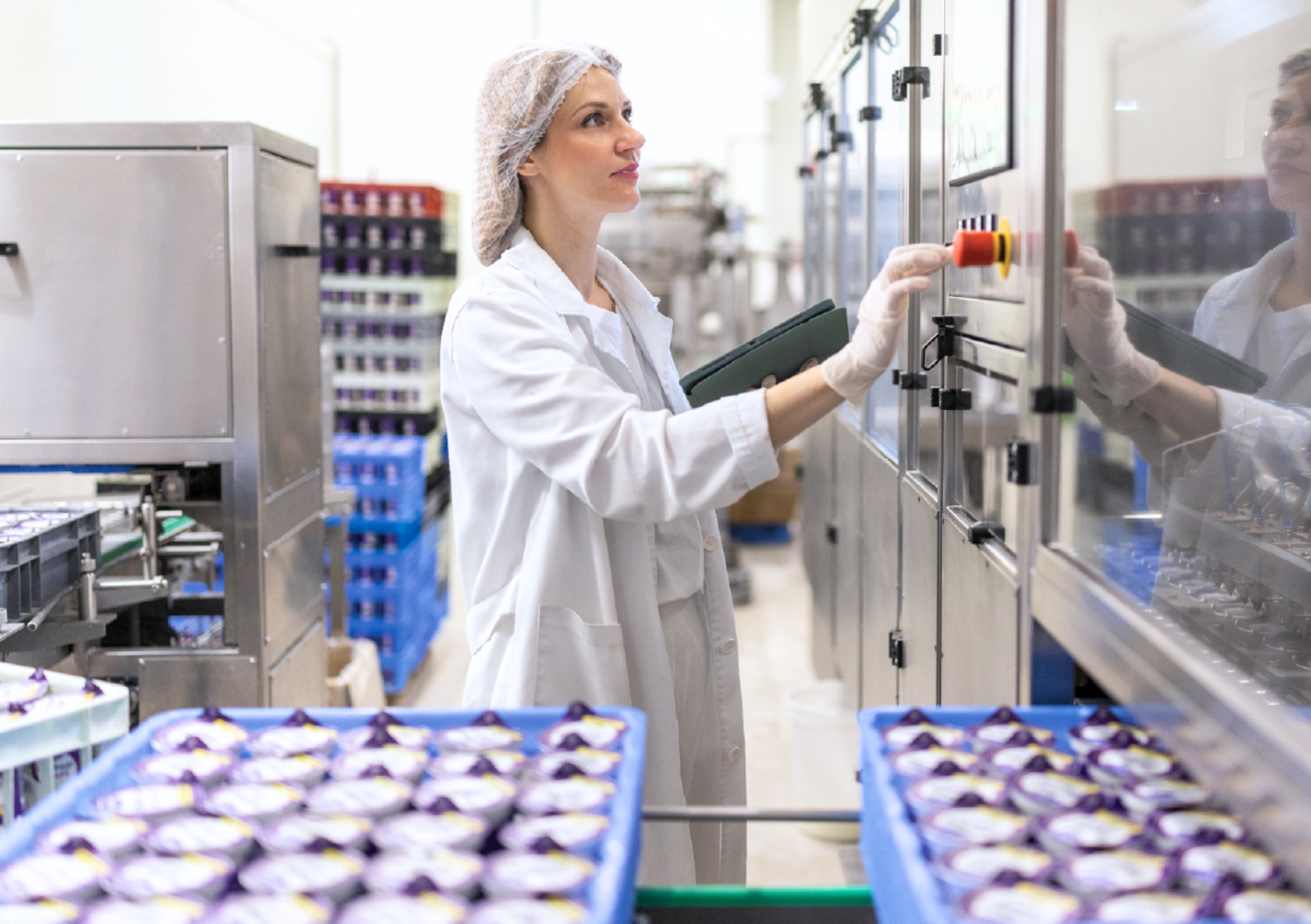 A woman in a white coat and hairnet operates a control panel in a factory. She stands near trays of packaged products, focusing intently.