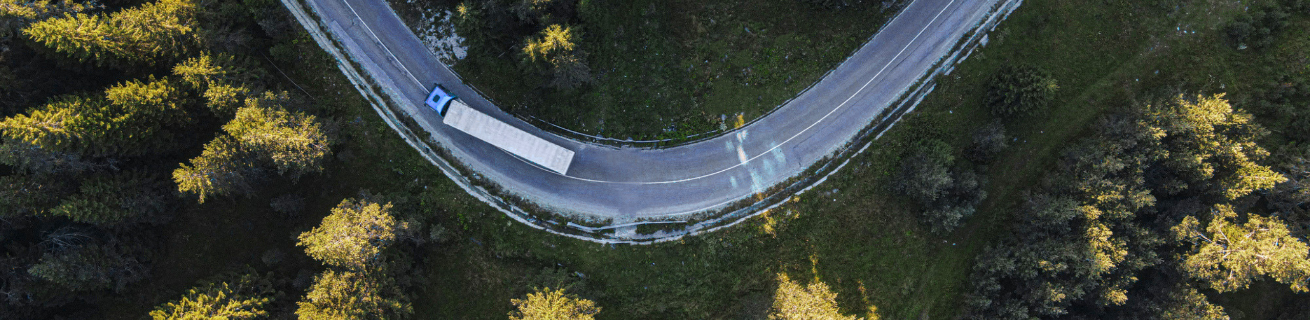 Aerial view of a curved road with a truck negotiating the turn, surrounded by trees. PAC.