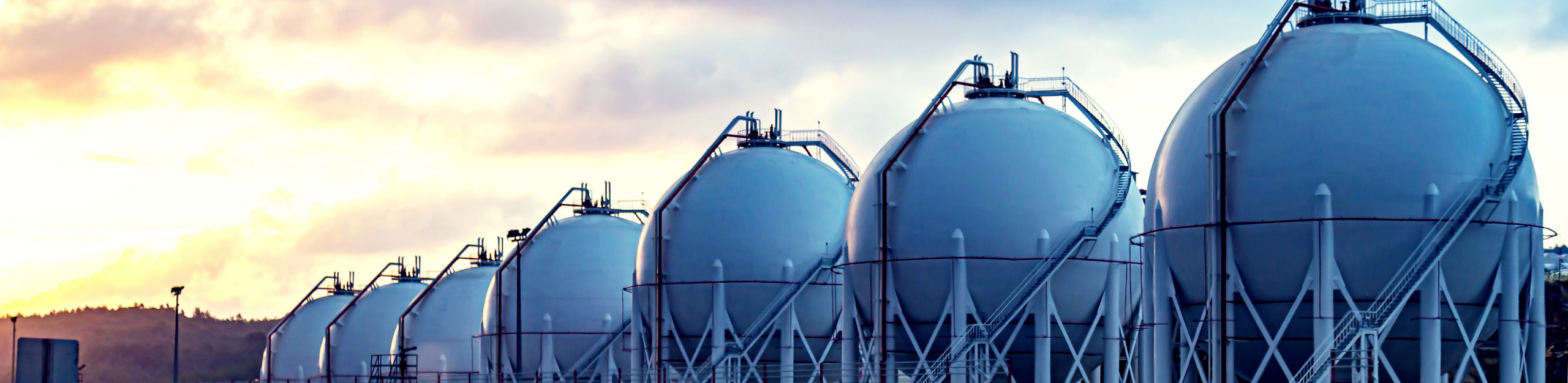 A row of large metal LNG tanks silhouetted against a vibrant sunset sky. LNG Analysis.