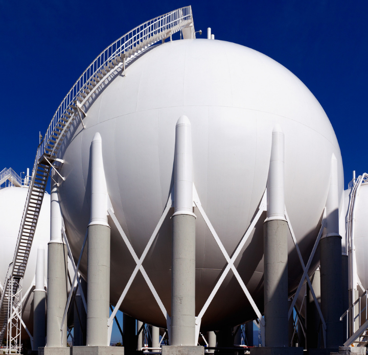 Large white storage tanks with a ladder on top, set against a clear blue sky. LNG Analysis.