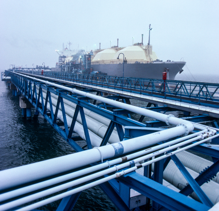 A large LNG ship rests at a pier, surrounded by water and with cargo containers stacked nearby. LNG Analysis.