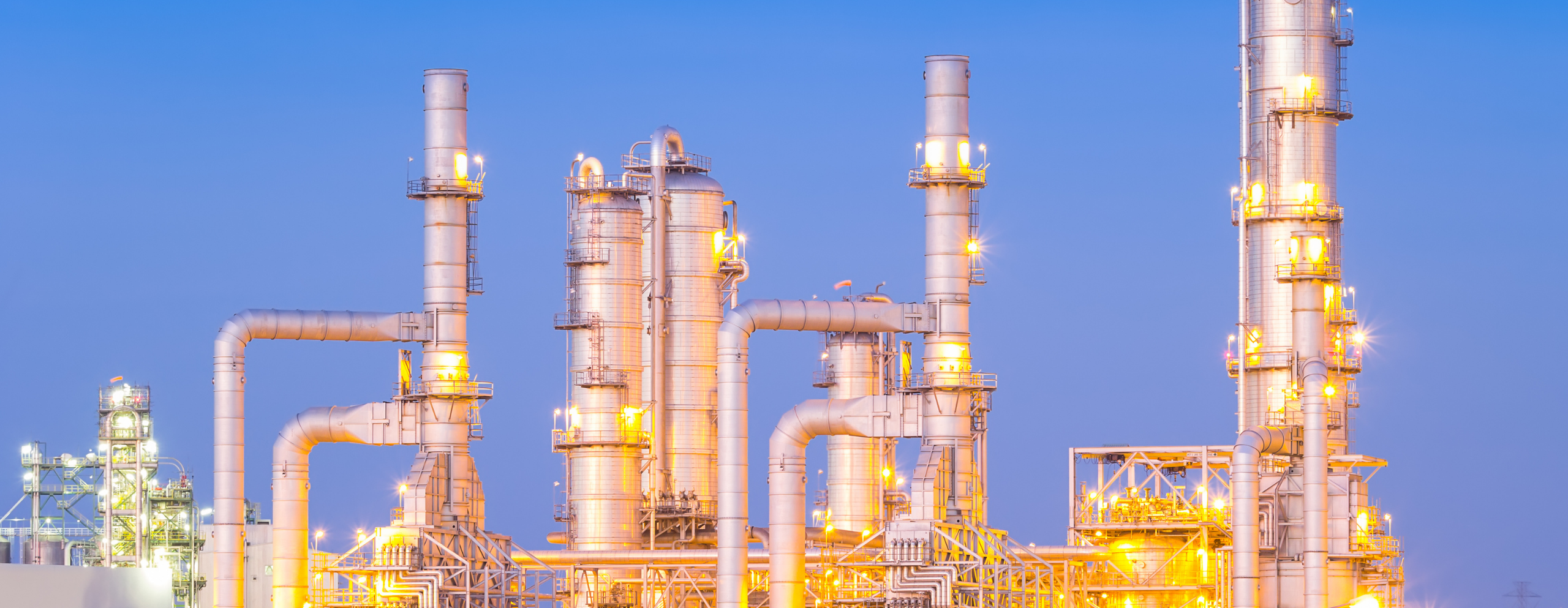 A large industrial refinery with towering smokestacks and complex machinery against a clear blue sky. LNG VAnalysis.